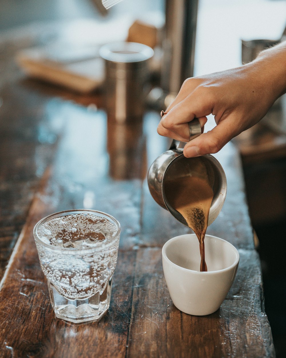 Coffee poured from a small steel jug into a ceramic cup on a worn timber bar