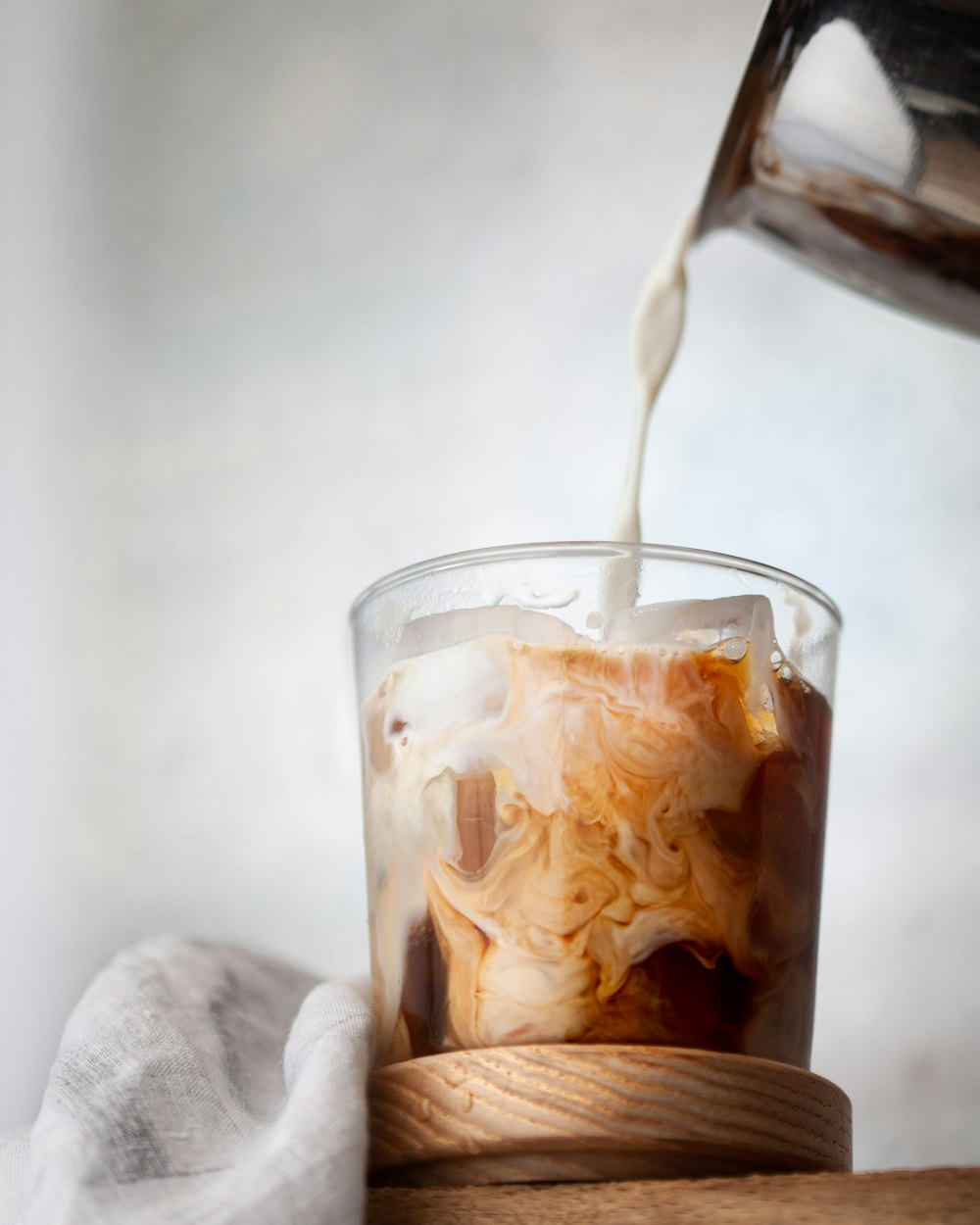Milk pouring into a tall glass of iced cold brew on a wooden coaster