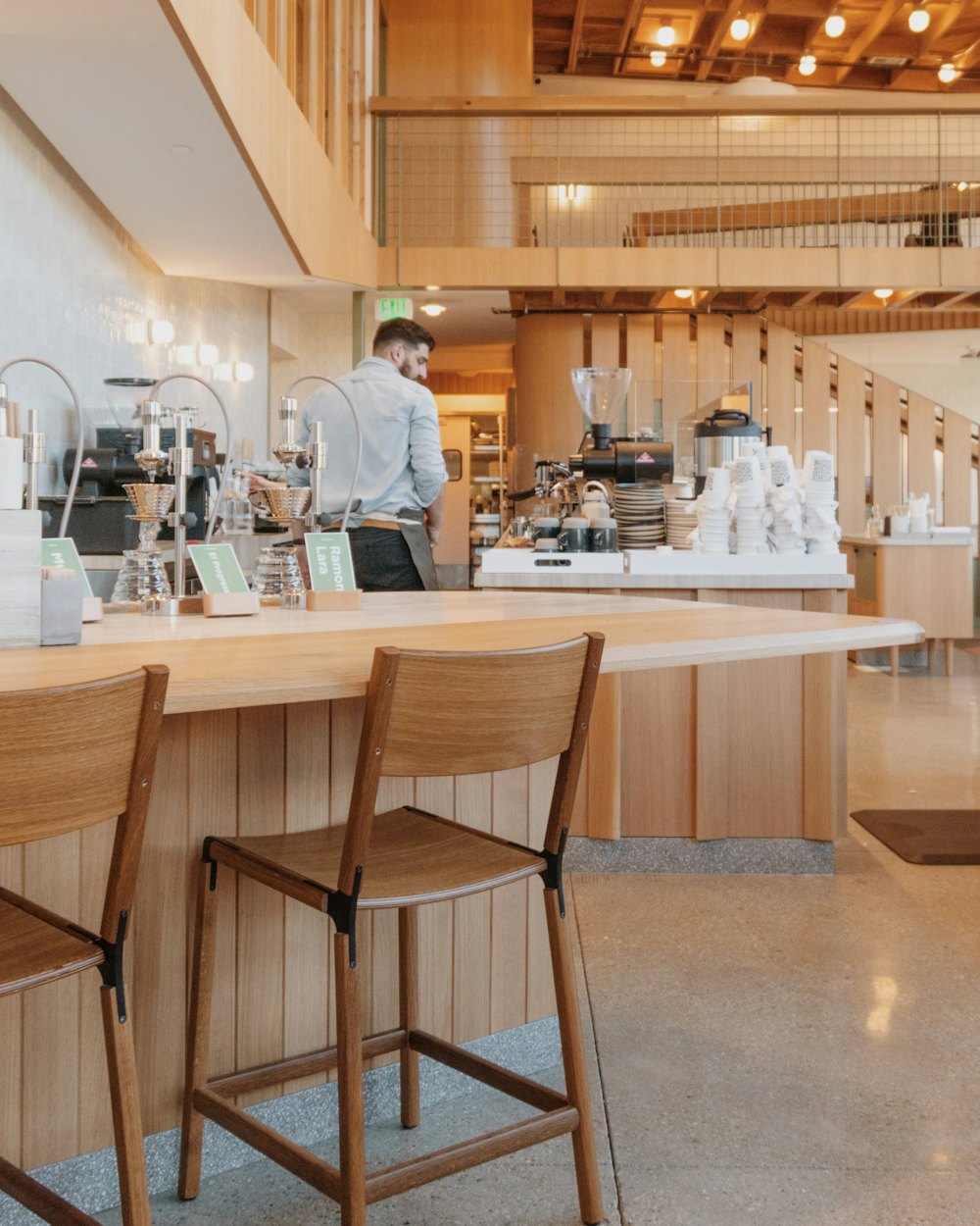 A barista working behind the pale timber espresso bar, cups stacked along the counter