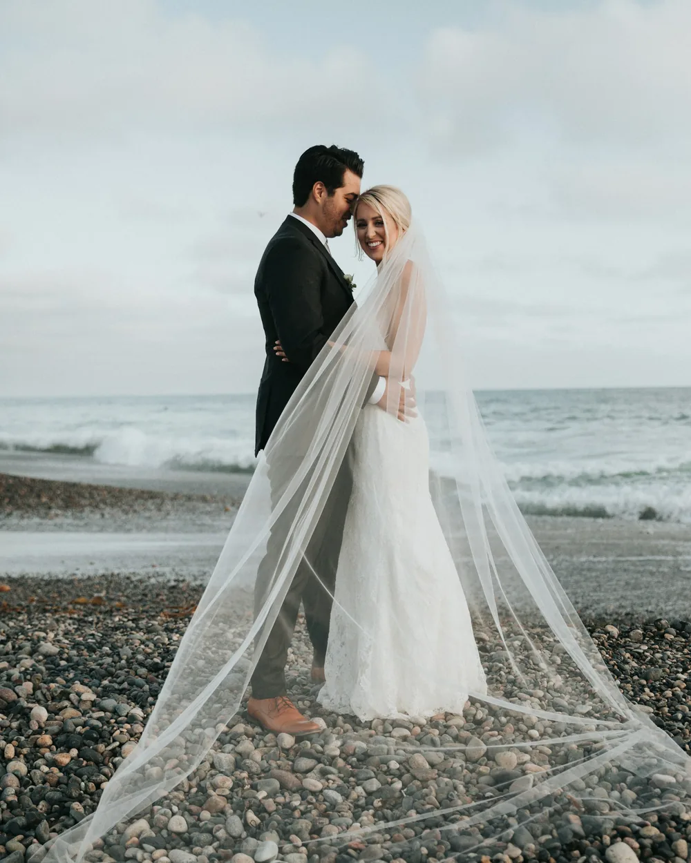 Bride and groom on a pebble beach, veil trailing in the wind