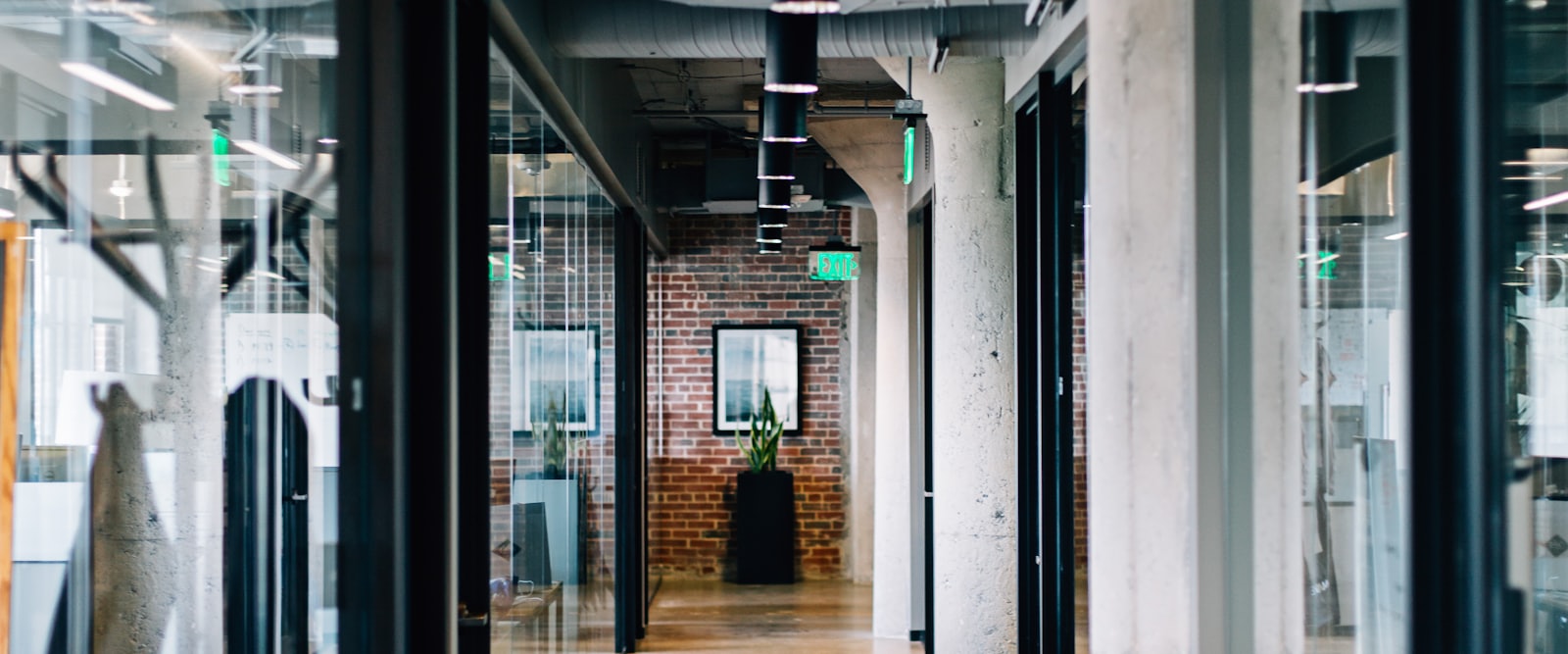 A long office corridor with glass partition walls, concrete columns and exposed ceiling ducts