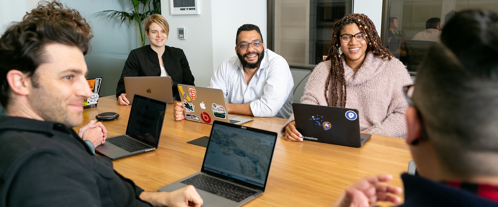 Four colleagues with open laptops around a meeting table, listening to a fifth person speaking